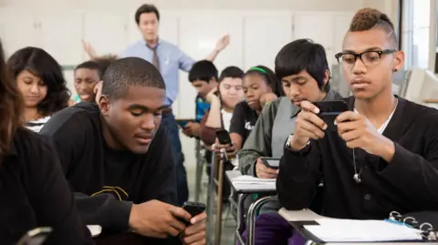 Getty Images Students sitting at desks in a classroom use cellphones as a teacher at the back of the classroom looks frustrated trying to get their attention